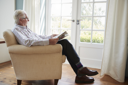Senior man sitting in an armchair reading newspaper at homeの写真素材