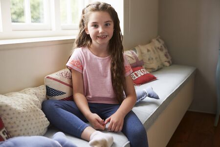 Portrait Of Smiling Young Girl Sitting On Window Seat At Homeの写真素材