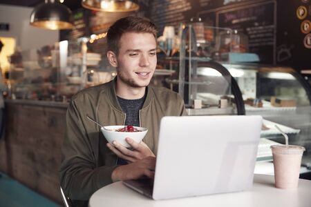 Young Man Using Laptop In Cafe Whilst Eating Breakfastの写真素材