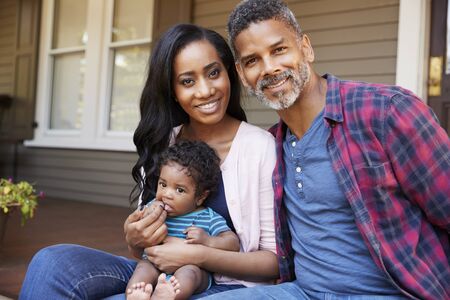 Family With Baby Son Sit On Steps Leading Up To Porch Of Home