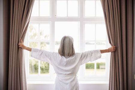 Senior Woman Opening Bedroom Curtains And Looking Out Of Windowの写真素材