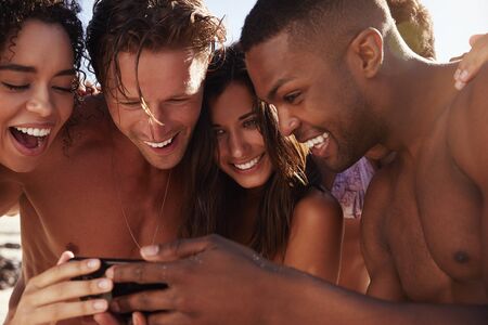 Friends On Beach Vacation Looking At Photo On Mobile Phoneの写真素材