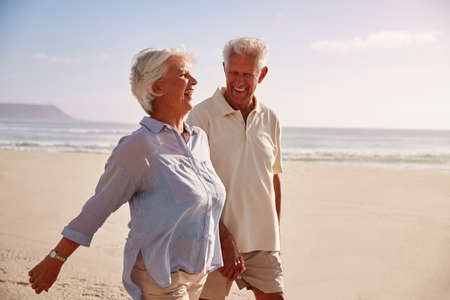 Senior Retired Couple Walking Along Beach Hand In Hand Together