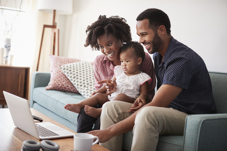 Family With Baby Daughter Sitting On Sofa At Home Looking At Laptop Computerの写真素材