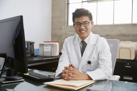Young Asian male doctor sitting at desk, portraitの写真素材