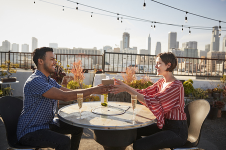 Man Giving Woman Gift As They Celebrate On Rooftop Terrace With City Skyline In Backgroundの写真素材