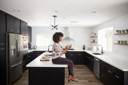 Woman At Home Sitting On Kitchen Island Whilst Using Mobile Phoneの写真素材