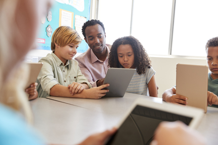 Teacher sits with young kids using computers in school classの写真素材