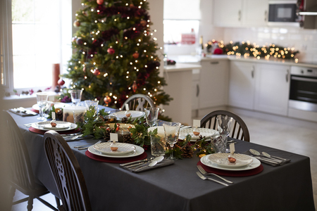 Christmas table setting with bauble name card holders arranged on plates and green and red decorations with Christmas tree and kitchen in the backgroundの写真素材
