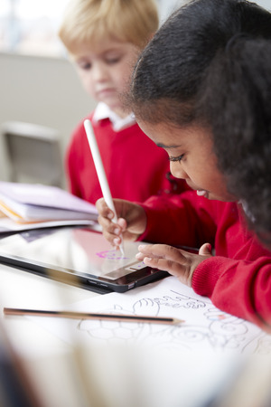Close up of a schoolgirl sitting at desk next to a boy in an infant school classroom, using a tablet computer and stylus, selective focus, verticalの写真素材