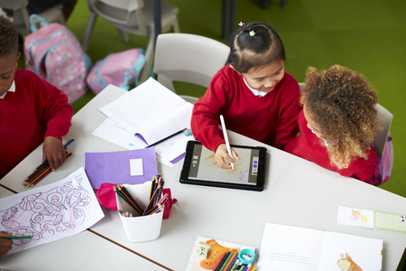 Elevated view of two infant school girls sitting at a table, using a tablet computer and stylus in a classroomの写真素材