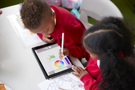 Elevated view of two kindergarten school kids sitting at a desk in a classroom drawing with a tablet computer and stylus, close upの写真素材