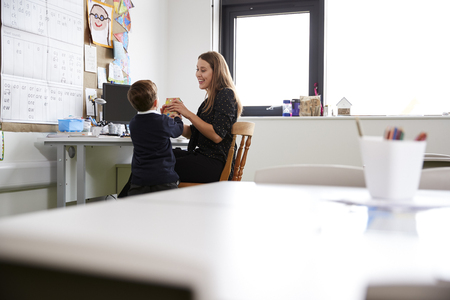 Schoolboy at primary school presenting a gift to his female teacher in a classroom, selective focus, back viewの写真素材