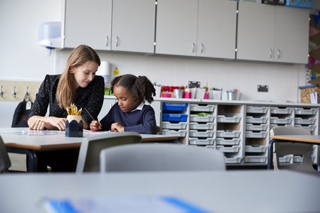 Young female primary school teacher working one on one with a schoolgirl sitting at table in a classroom, selective focusの写真素材
