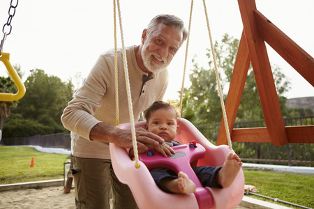Senior Hispanic grandfather pushing his baby grandson on a swing at a playground in the parkの写真素材