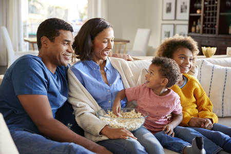Toddler boy eating popcorn, sitting on the sofa with his sister and parents in their living room...