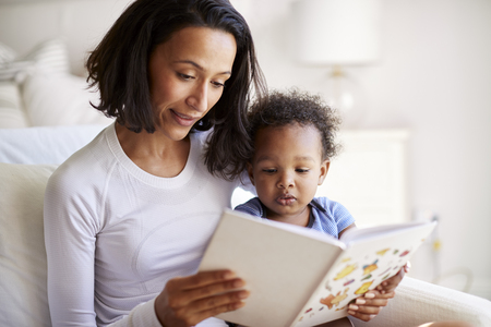 Close up of mixed race young adult mother sitting in an armchair reading a book with her two year old son, close upの写真素材