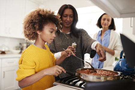 Pre-teen black girl standing at the hob in the kitchen preparing food with her grandmother and mother, close up, selective focusの写真素材
