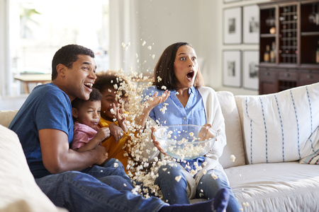 Young mixed race family sitting together on the sofa in their living room watching a scary movie accidentally throwing popcorn in the airの写真素材