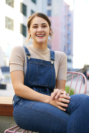 Trendy young Hispanic woman wearing dungarees sitting in the street laughing to camera, close upの写真素材
