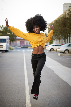 Young black woman with afro jumps, looking to camera, while crossing street, front view, verticalの写真素材