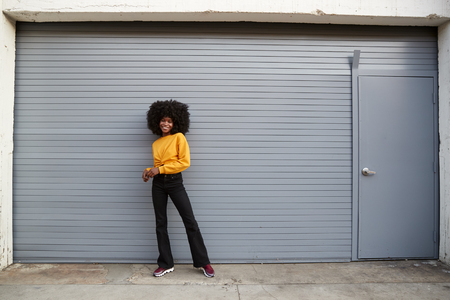 Hip young black woman standing against grey security shutters smiling to camera, full lengthの写真素材