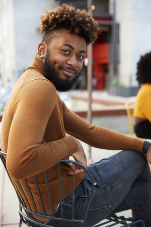 Millennial black hipster man sitting outside a cafe in the street, turns smiling to camera, close upの写真素材