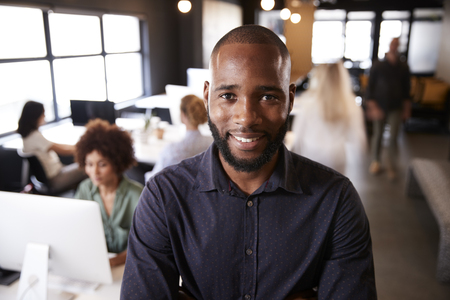Bearded black male creative standing in a busy casual office, smiling to cameraの写真素材