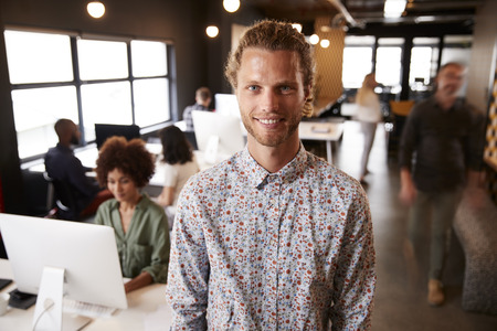 Millennial white male creative standing in a busy casual office, smiling to cameraの写真素材