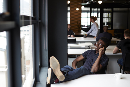 Young black male creative sitting in an office with his feet up on the desk using phone, close upの写真素材
