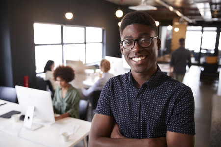 Millennial black male creative standing in a busy casual office, smiling to cameraの写真素材