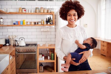 Portrait Of Smiling Mother Holding Sleeping Baby Son In Kitchenの写真素材