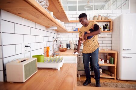 Multi-Tasking Father Holds Sleeping Baby Son And Cleans In Kitchenの写真素材