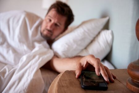 Mid adult man lying in bed, reaching out to smartphone on the bedside table in the foregroundの写真素材