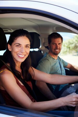Mid adult white woman driving car, husband in front passenger seat smiling to camera, vertical
