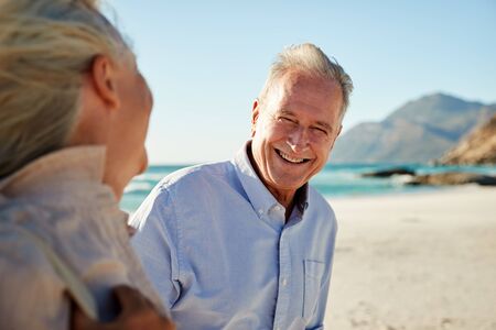 Senior white couple walking on a beach, looking at each other and smiling, waist up, close up
