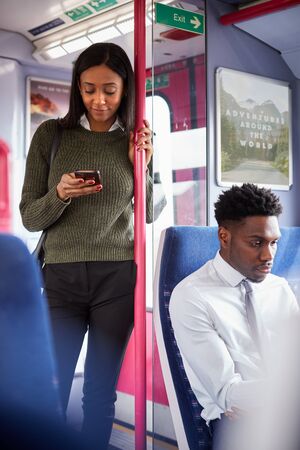 Female Passenger Standing By Doors In Train Looking At Mobile Phone