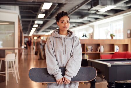 Portrait Of Casually Dressed Young Businesswoman With Skateboard Standing In Open Plan Workplaceの写真素材
