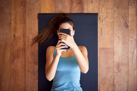 Overhead View Of Woman Lying On Exercise Mat Wearing Wireless Earphones Connected To Mobile Phoneの写真素材