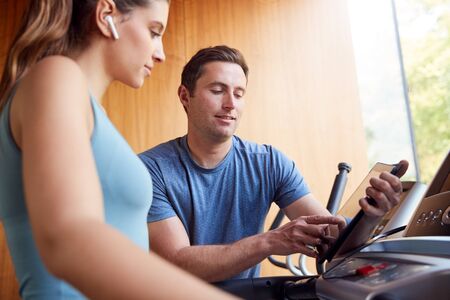 Woman In Gym With Personal Trainer Analysing Performance Using Smart Watch And Digital Tablet