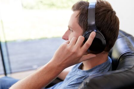 Man Relaxing In Chair At Home Listening To Music Or Podcast Music On Wireless Earphones
