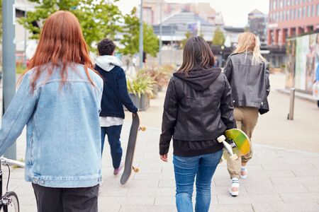 Rear View Of Female Friends With Skateboards And Bike Walking Through City Centerの写真素材