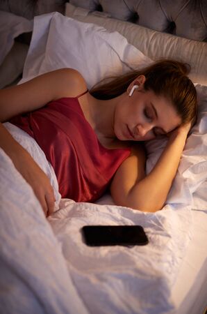 Woman Sleeping In Bed Wearing Wireless Earphones Connected To Mobile Phoneの写真素材