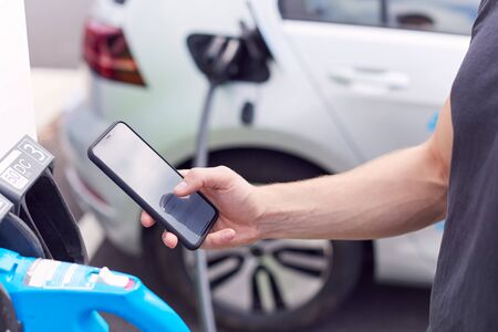 Man Charging Electric Vehicle With Cable Looking At App On Mobile Phone