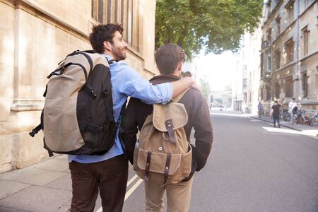 Rear View Of Male Gay Couple On Vacation Wearing Backpacks Walking Along City Streetの写真素材