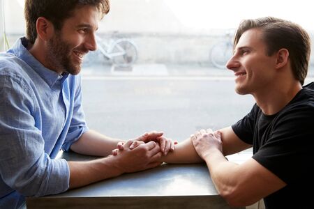 Loving Male Gay Couple Sitting At Table In Coffee Shop Holding Handsの写真素材