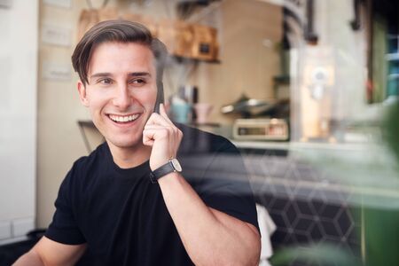 Young Man In Coffee Shop Talking On Mobile Phone Viewed Through Window