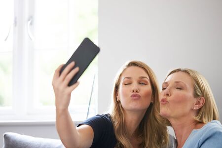 Senior Mother With Adult Daughter Sitting On Sofa Posing For Selfie On Mobile Phone