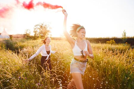 Two Female Friends Camping At Music Festival Running Through Field With Smoke Flareの写真素材
