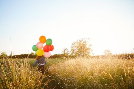 Rear View Of Two Female Friends Camping Running Through Field With Balloons Against Flaring Sunの写真素材